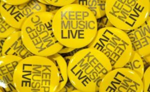 Image of a pile of yellow circular badges with the words "Keep Music Live"