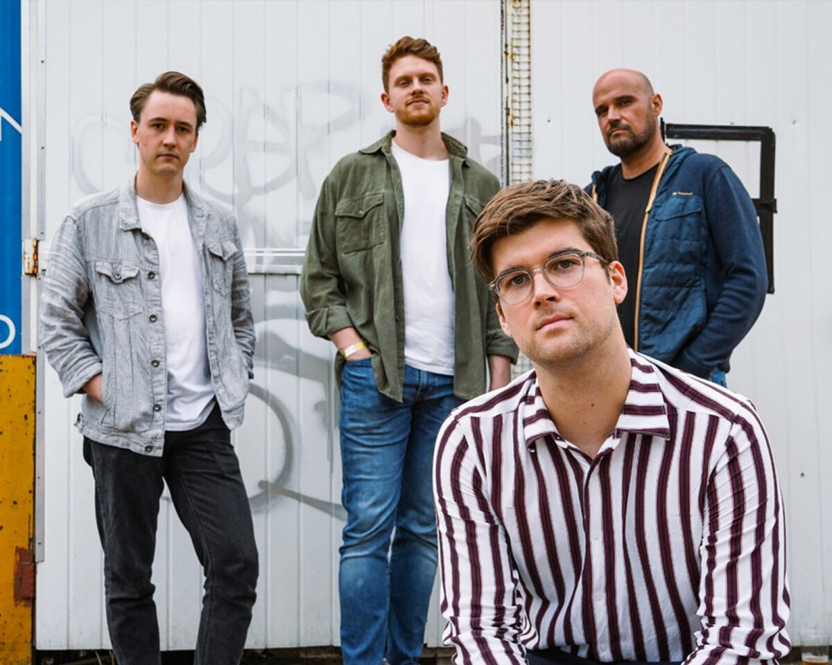 a four-person band posing against a white shipping container or metal wall. In the foreground sits a man wearing glasses and a burgundy and white vertically striped shirt. Standing behind him are three other men: on the left, a man in a light grey/striped jacket over a white t-shirt with dark trousers; in the middle, a man with reddish/ginger hair wearing a green/olive overshirt with a white t-shirt and blue jeans; and on the right, a man with a shaved head in a navy blue jacket or hoodie. All four have serious expressions common in band promotional photos. The setting appears industrial, with the clean white background providing contrast to their casual but coordinated styling.