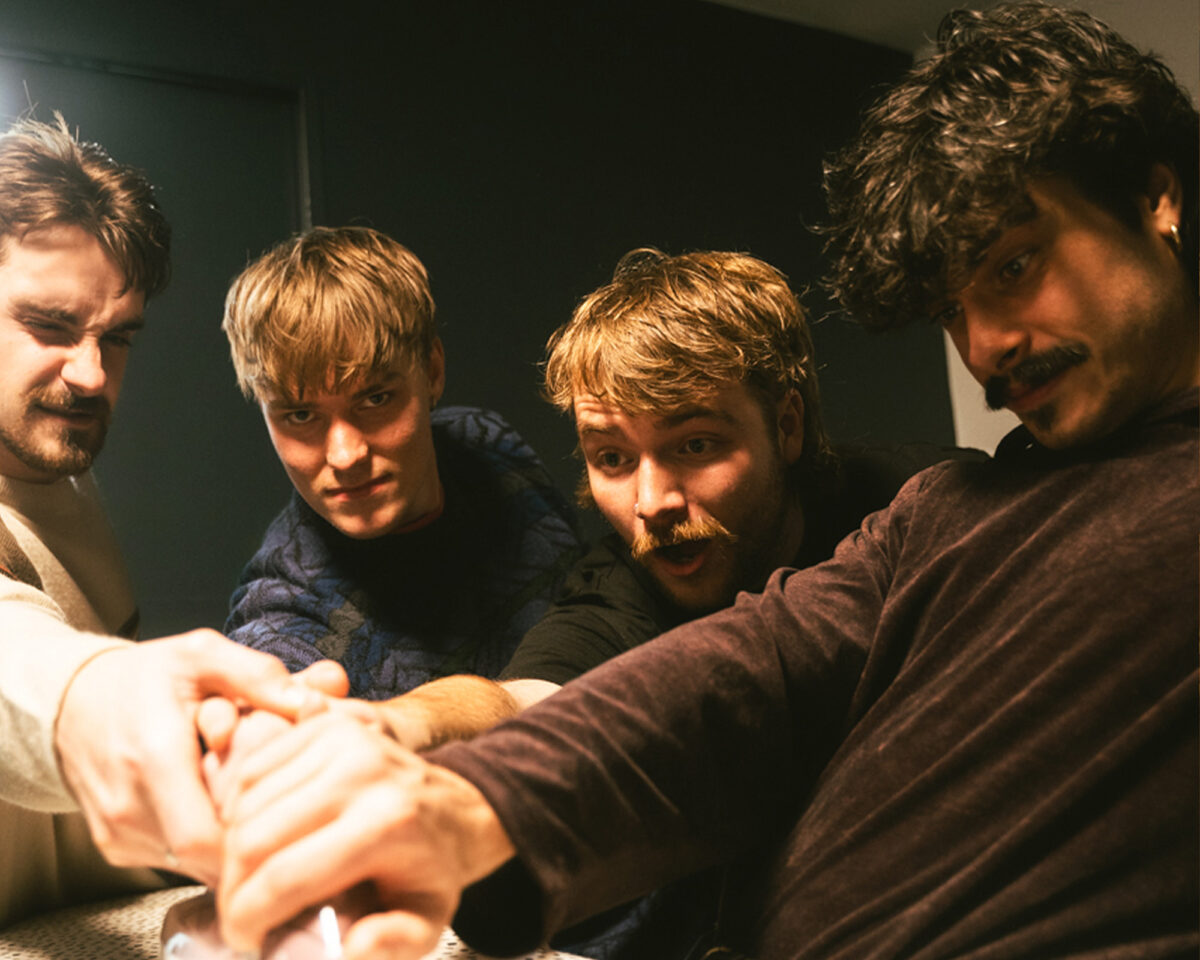The image shows four young men with their hands stacked together in what appears to be a team huddle or solidarity gesture. They all have their faces turned toward the camera while their hands are joined in the foreground of the image.