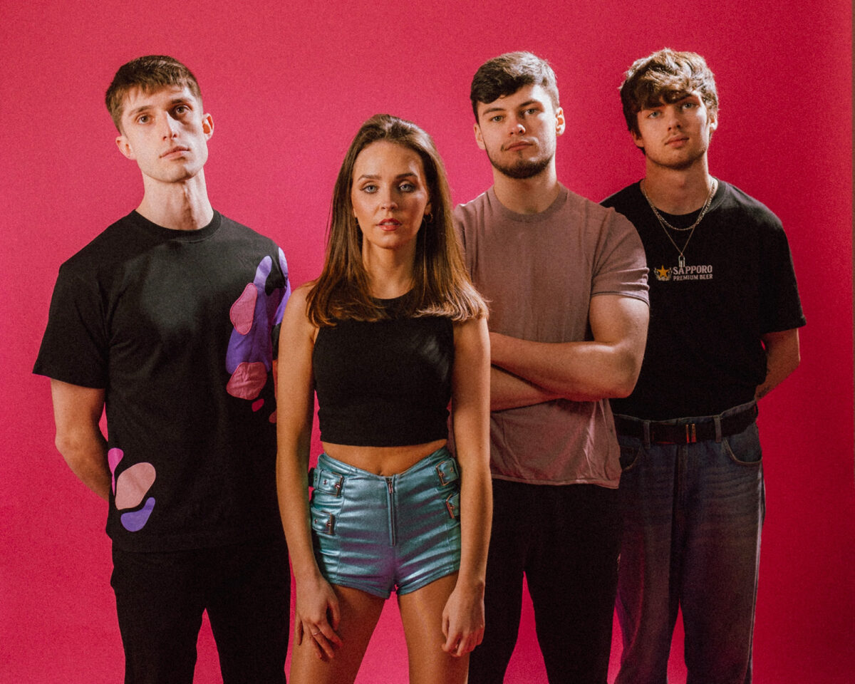 A four-person music group posing against a bright pink/red background. The group consists of three men and one woman arranged in a row.