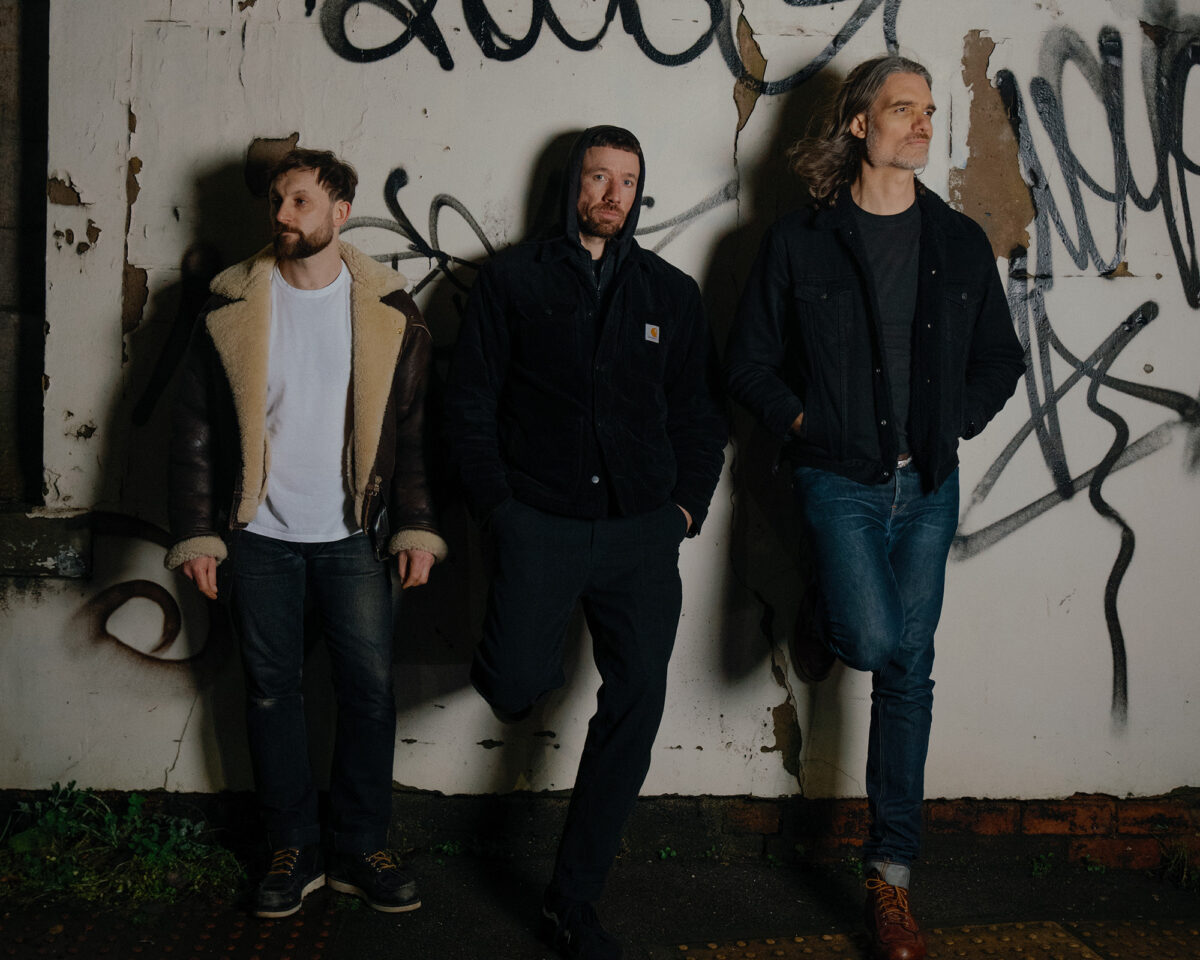 Three men standing against a white wall covered with graffiti and peeling paint. They appear to be a band or music group in a promotional photo.