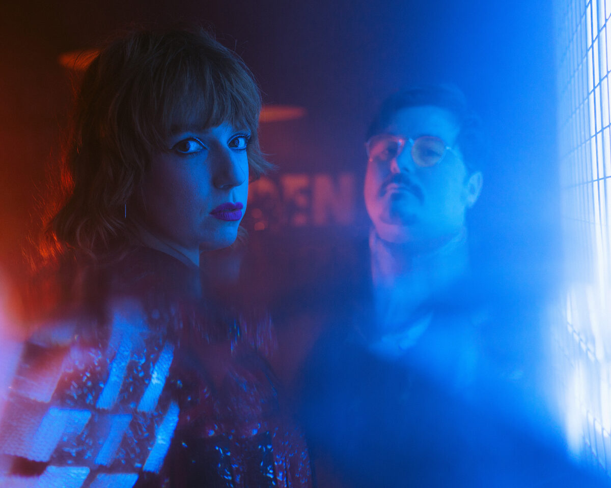 two people in a moody, atmospheric setting with dramatic red and blue lighting. On the left is a person with blonde hair and bangs looking directly at the camera. On the right, partially obscured and more blurred, is a person with glasses and what appears to be facial hair.