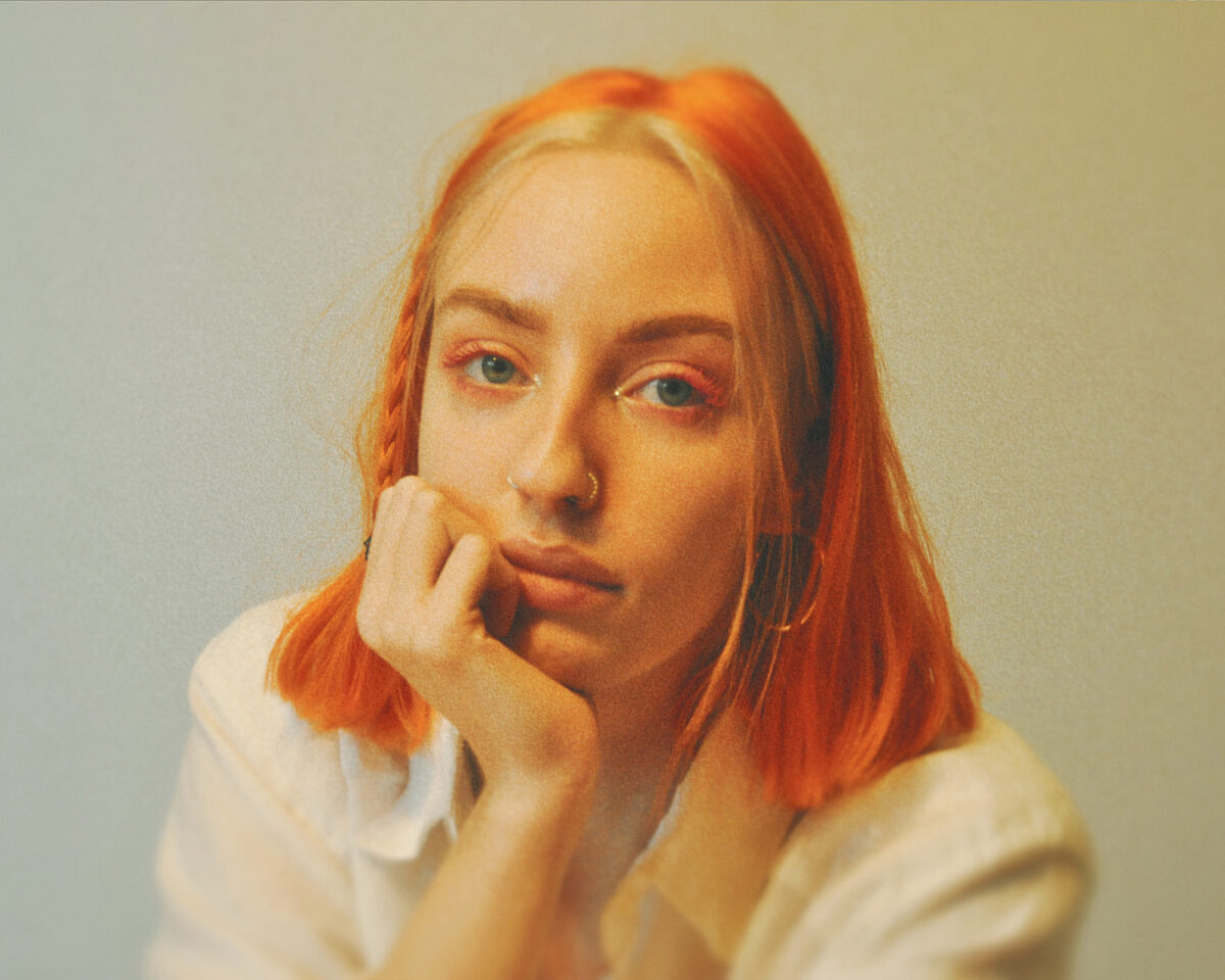 a close-up portrait of a person with bright orange/peachy hair cut to about chin length. They have light blue eyes with matching orange/peachy eyeshadow and are wearing a white or cream-coloured top. The person has a thoughtful expression with their hand resting against their cheek, looking directly at the camera. They appear to have a small nose piercing. The photo has a soft, slightly vintage aesthetic with warm tones and a gentle film-like grain overlay. The background is a neutral light colour, putting the focus entirely on the subject. The overall mood is contemplative and serene.