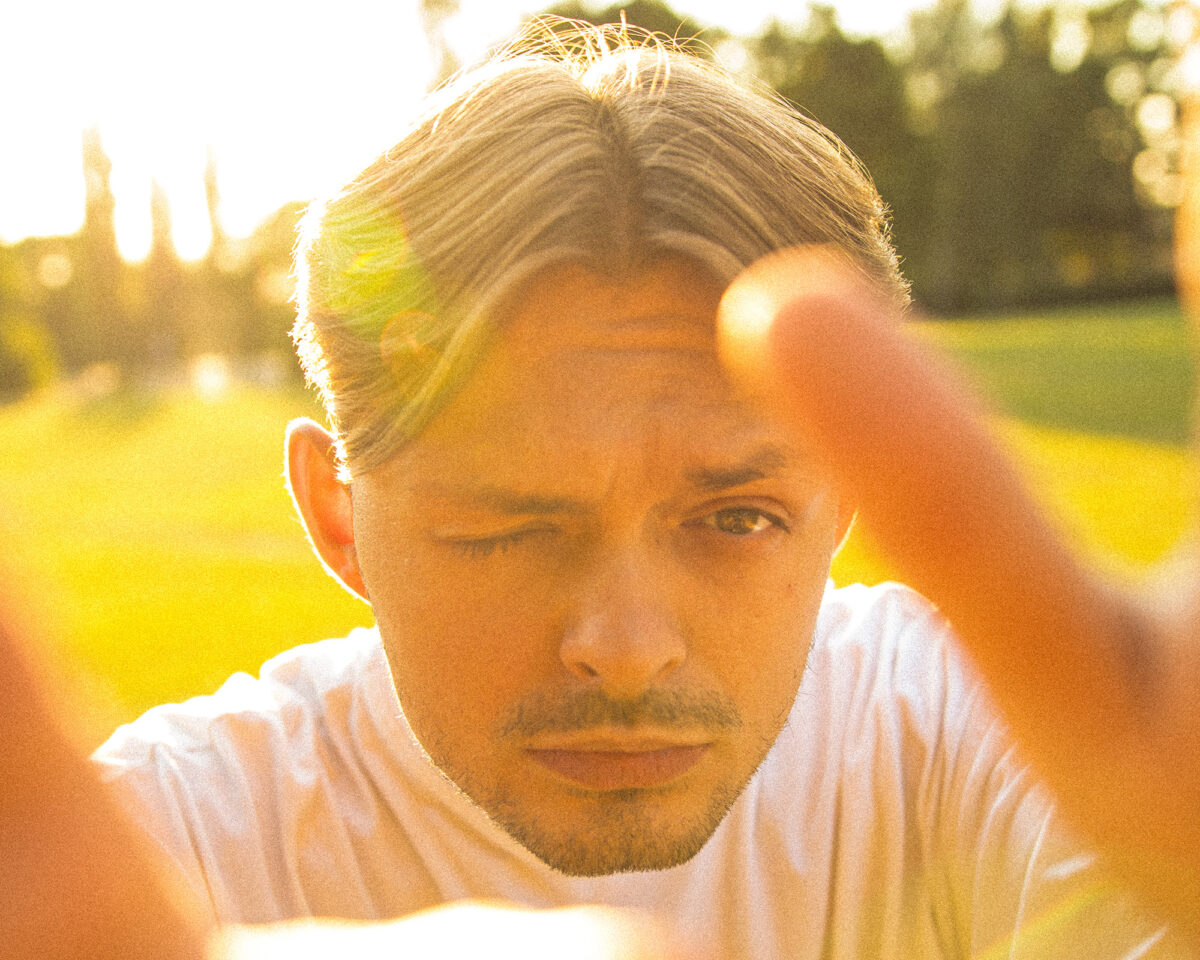 a close-up portrait of a young man with short, neatly parted hair, wearing a white t-shirt. He has a thoughtful expression with one eye closed or squinting and facial hair along his jawline and upper lip.