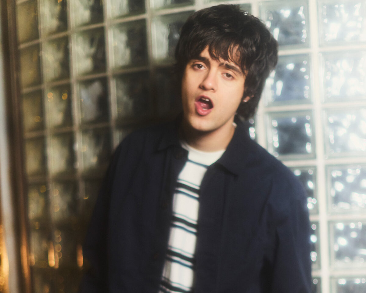The image shows a young person with dark, shaggy hair, wearing a dark jacket over a white and dark striped shirt. They appear to be singing or speaking with their mouth open, captured in a portrait-style photograph. The background features a glass block wall that allows light to filter through, creating a soft, atmospheric effect. The lighting gives the image a slightly warm tone on one side while maintaining cooler tones elsewhere. The subject is positioned slightly off-center in the frame, with their face clearly visible against the textured background.