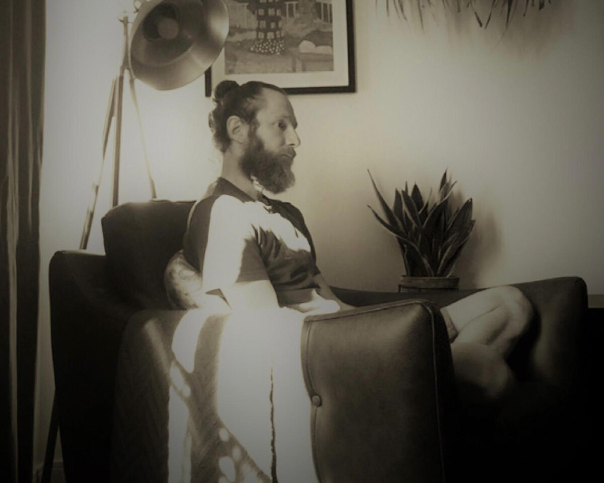 a black and white photograph of a person with a beard and hair in a bun sitting in what appears to be a living room. They're seated on a dark sofa or armchair, wearing what looks like a light-coloured top with dark elements (possibly overalls or a vest).