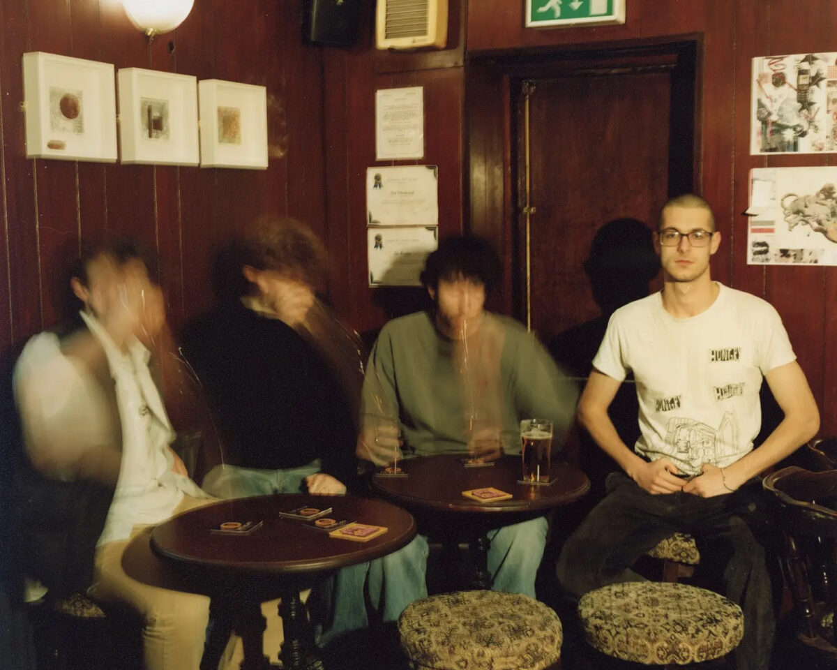 The image shows four people sitting at small round tables in what appears to be a pub or bar with dark wood-panelled walls. Three of the people on the left side are captured in motion blur, suggesting they were moving when the photo was taken. The person on the right is clearly visible - a young man with glasses wearing a white t-shirt with some text and graphics on it.