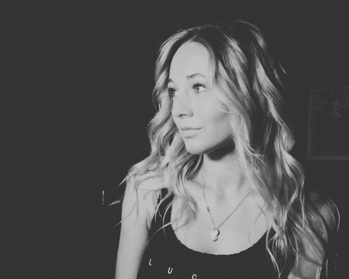 a person with long, wavy hair looking off to the side against a dark background. They are wearing a dark tank top or camisole with the word "LUCKY" printed on it. A delicate necklace with a small pendant can be seen around their neck. The portrait has a dramatic, high-contrast look with the subject illuminated against the predominantly dark background. The person has a thoughtful expression and their hair appears to have lighter highlights that stand out in the monochrome image. The composition is in a square format with the subject positioned slightly to the right of the frame.