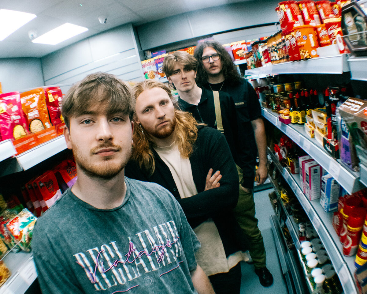 four young men standing in a narrow convenience store aisle, arranged in a line from foreground to background. They appear to be a band or music group posing for a promotional photo. The person in the foreground is wearing a grey t-shirt with "PROHIBITION" text visible. Behind him is someone with longer reddish-blonde hair, followed by a person with brown hair, and in the back, someone with longer dark curly hair and glasses.