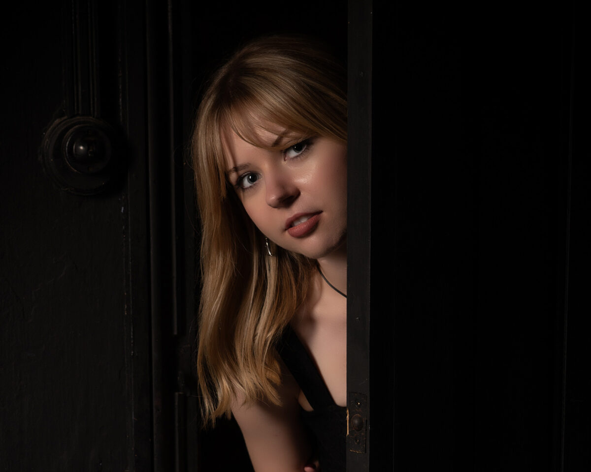 a person with long blonde hair and bangs peering through a partially opened dark door. The photograph has a dramatic, moody aesthetic with low-key lighting that creates strong contrast between the dark door and the illuminated face. The person has a somewhat concerned or contemplative expression and appears to be wearing dark clothing. They are looking directly at the camera, visible only from the small opening in the doorway. The overall composition creates an atmosphere of mystery or intrigue, with most of the frame filled by the dark door and surrounding darkness, making the illuminated face the focal point of the image.