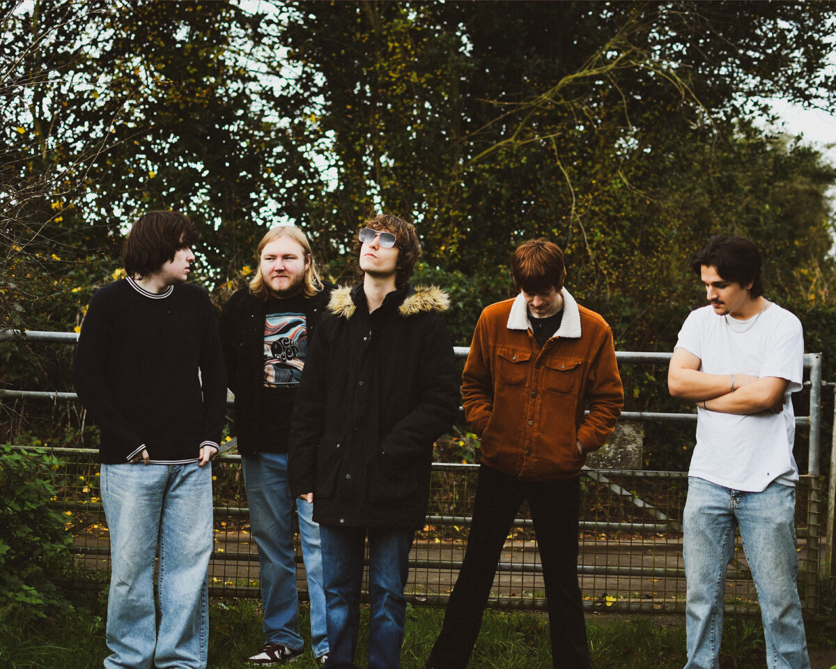 five young men standing in a line against a metal railing fence with trees and foliage in the background. They appear to be a music group or band posing for a promotional photo. From left to right: A person with dark hair wearing a black top with striped trim and light blue jeans A person with longer blonde/light brown hair and a beard wearing a graphic t-shirt and jeans A person in the middle wearing sunglasses and a black coat with fur-trimmed hood A person wearing a brown corduroy or suede jacket with sherpa/fleece collar A person on the right in a white t-shirt with arms crossed, wearing light blue jeans The setting appears to be outdoors in a park or natural area