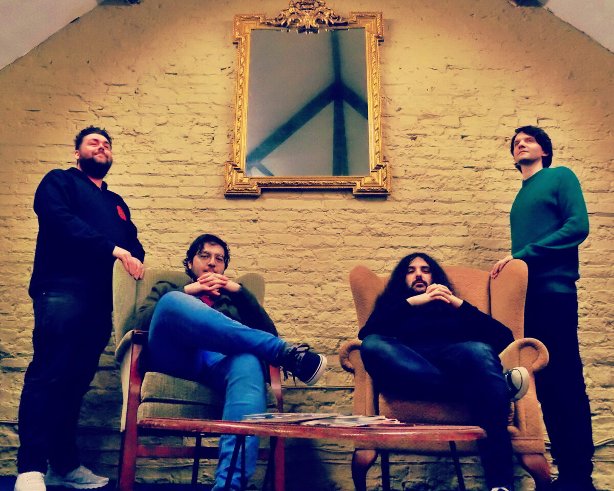 four men arranged in what appears to be a promotional photo for a band. They're positioned in a room with cream-coloured painted brick walls and a distinctive ornate gold-framed mirror hanging centrally on the wall.