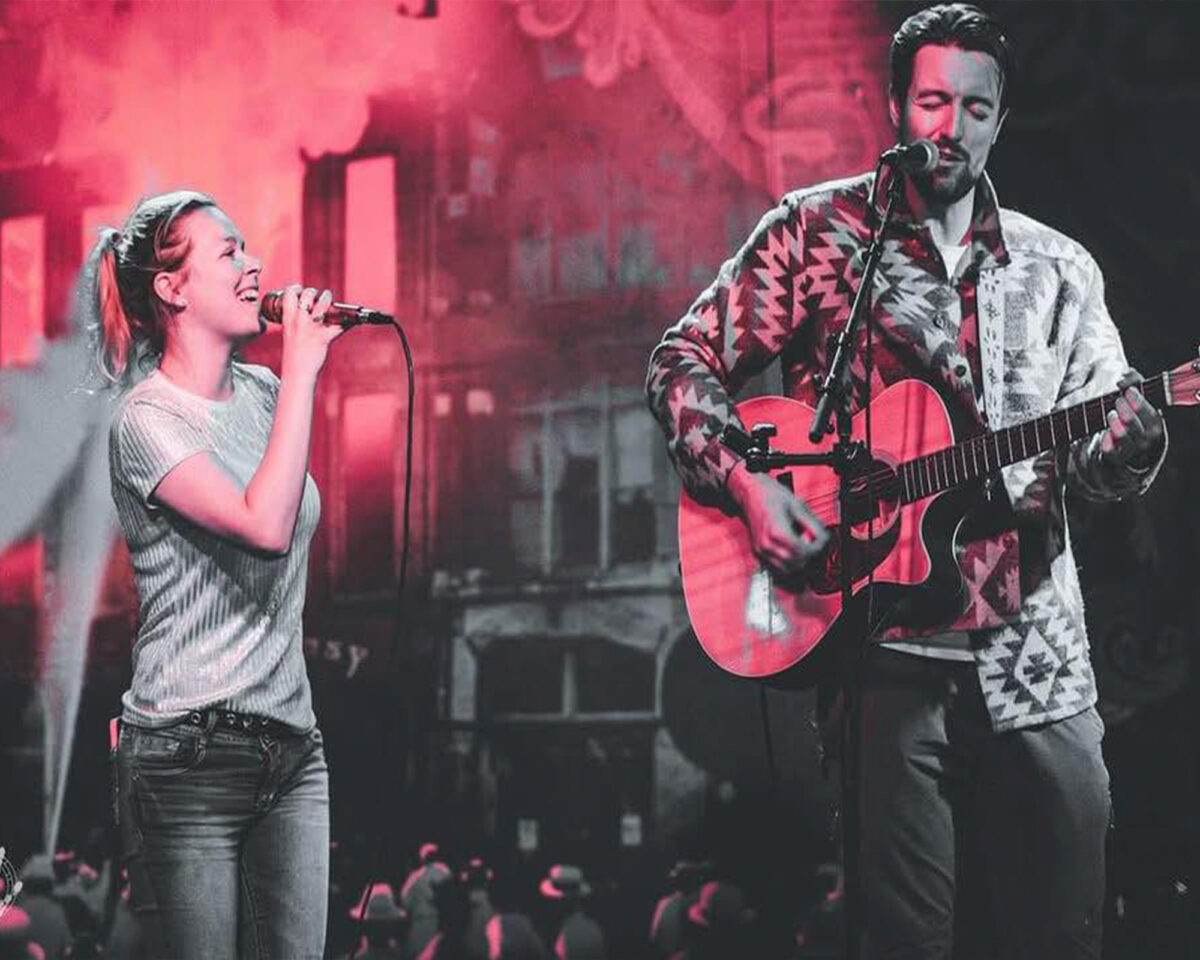 Two musicians performing on stage together. On the left is a woman with her hair in a ponytail, wearing a light-coloured striped or ribbed top and jeans, singing into a microphone. On the right is a man with facial hair wearing a patterned cardigan or jacket with geometric designs, playing an acoustic guitar and singing into his own microphone.