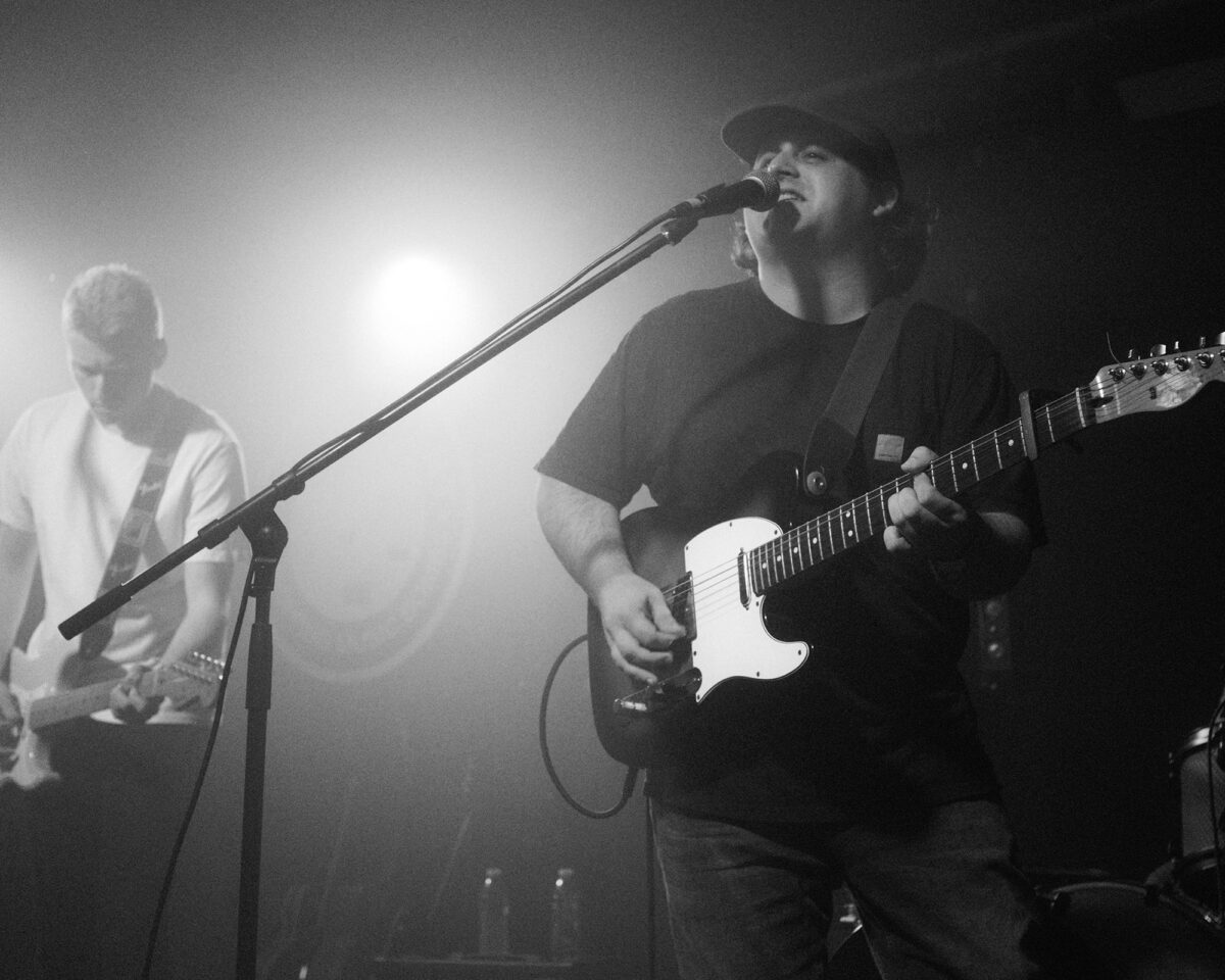 musicians performing on a foggy, atmospherically lit stage. In the foreground on the right is a guitarist wearing a cap and dark t-shirt, singing into a microphone while playing what appears to be a Telecaster-style electric guitar with a white pickguard. To the left and slightly behind him is another musician in a lighter coloured top, also playing guitar but less prominently featured and partially obscured by stage fog.