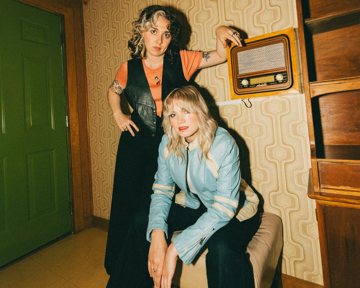 Two people in a vintage-styled room with geometric wallpaper. One person is standing, wearing an orange shirt under a black vest/waistcoat, with curly hair and visible tattoos on their arm. They're leaning against a vintage radio that's mounted on the wall.