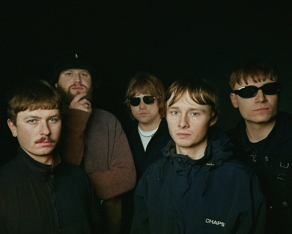 a group of five men arranged together for what appears to be a band photo against a plain black background. In the foreground is a young man with a blue jacket that has "CHAPS" visible on it. To his left is a man with a thin moustache. Behind them are three other band members - one wearing a black cap and brown jumper, and two wearing sunglasses. The overall aesthetic has a slightly retro feel, with some of the men sporting styling reminiscent of 1990s or early 2000s indie music scenes, including the distinctive haircuts and facial hair. The lighting creates a somewhat moody atmosphere while keeping their faces clearly visible against the dark backdrop. Their serious expressions and positioning suggest this is a promotional photograph for a musical group.