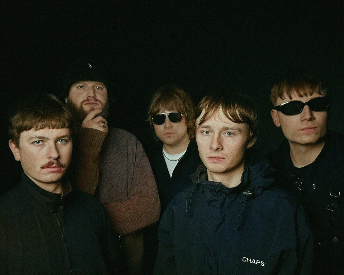 a group of five men arranged together for what appears to be a band photo against a plain black background. In the foreground is a young man with a blue jacket that has "CHAPS" visible on it. To his left is a man with a thin moustache. Behind them are three other band members - one wearing a black cap and brown jumper, and two wearing sunglasses. The overall aesthetic has a slightly retro feel, with some of the men sporting styling reminiscent of 1990s or early 2000s indie music scenes, including the distinctive haircuts and facial hair. The lighting creates a somewhat moody atmosphere while keeping their faces clearly visible against the dark backdrop. Their serious expressions and positioning suggest this is a promotional photograph for a musical group.
