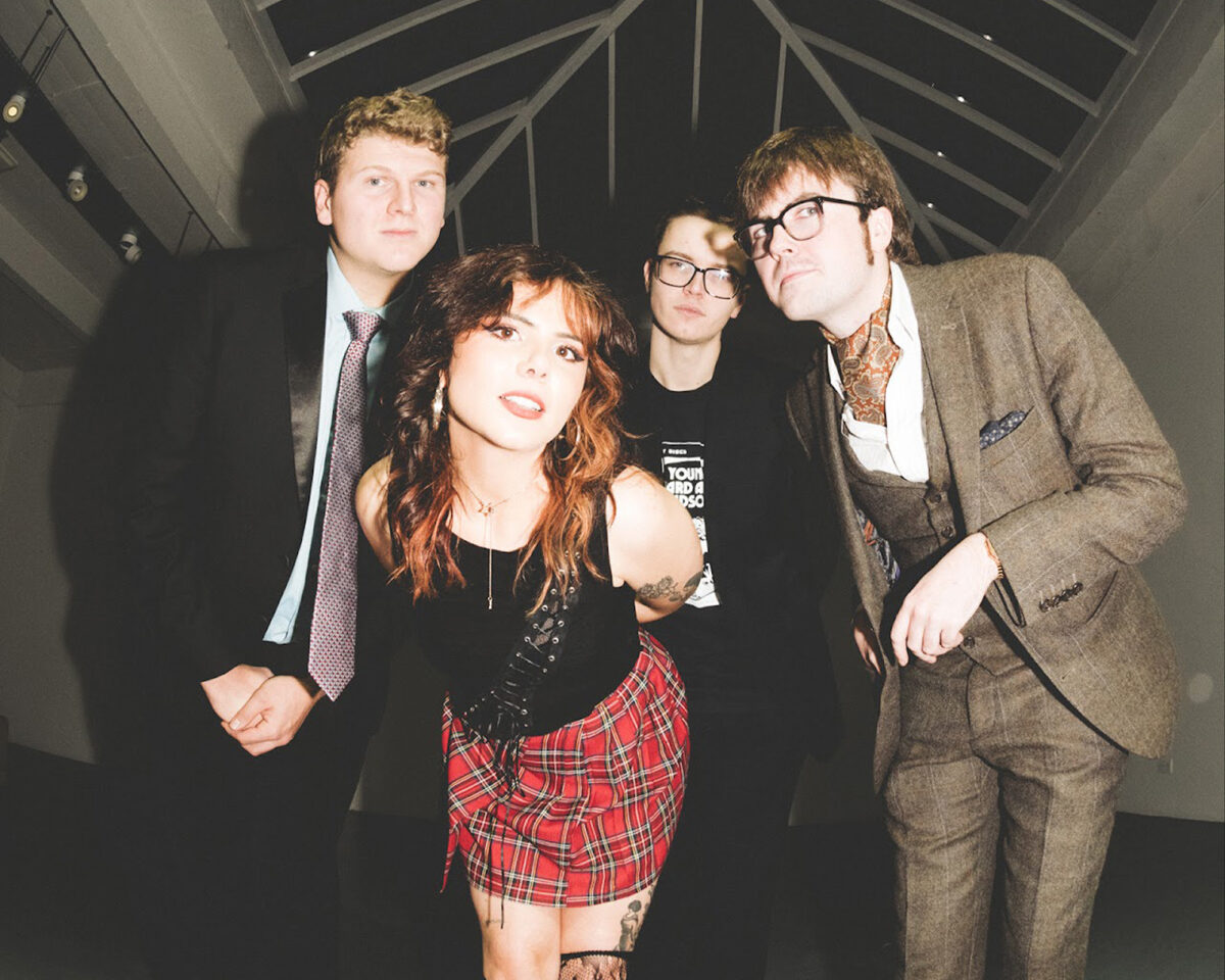 a group of four people posed together, likely a band, under what appears to be a glass or conservatory-style ceiling.