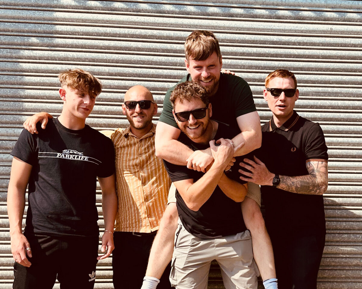 Five men posing together in front of a metallic roller shutter or garage door. They appear to be a band or music group taking a promotional photo. The group is arranged with four men standing in a row, while a fifth man (wearing a dark green t-shirt) is playfully hugging or climbing on the back of one of them (who is wearing sunglasses and light-coloured shorts).
