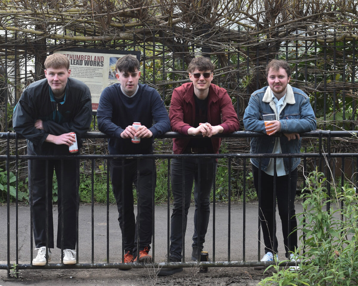 Four young men standing in a row along a metal railing or fence, each holding what appears to be a drink can or cup. Behind them is a sign that partially reads "THUMBERLAND" and some kind of natural structure with branches or vines.