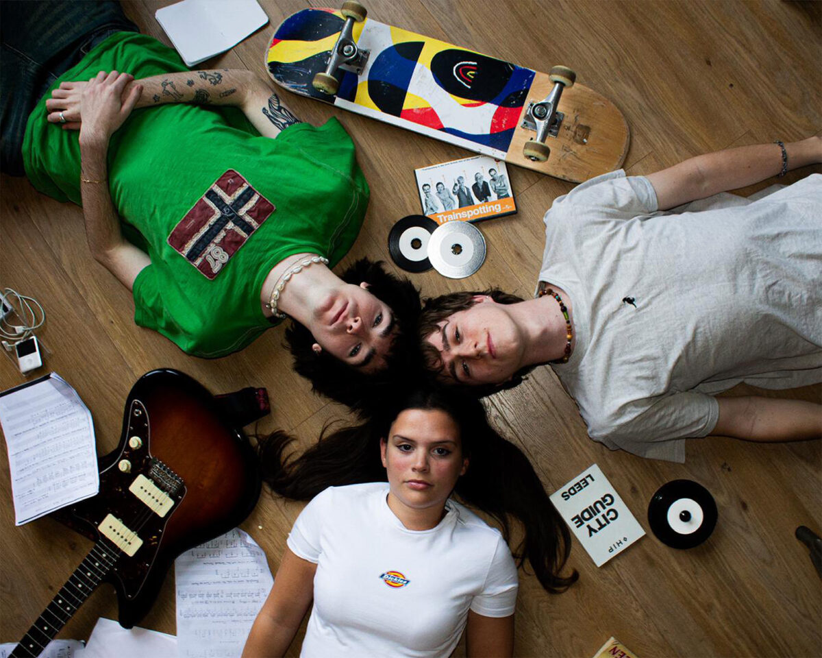 Three young people lying on a wooden floor with their heads together in the center, creating a star-like formation. They're surrounded by various objects related to music and skateboarding.