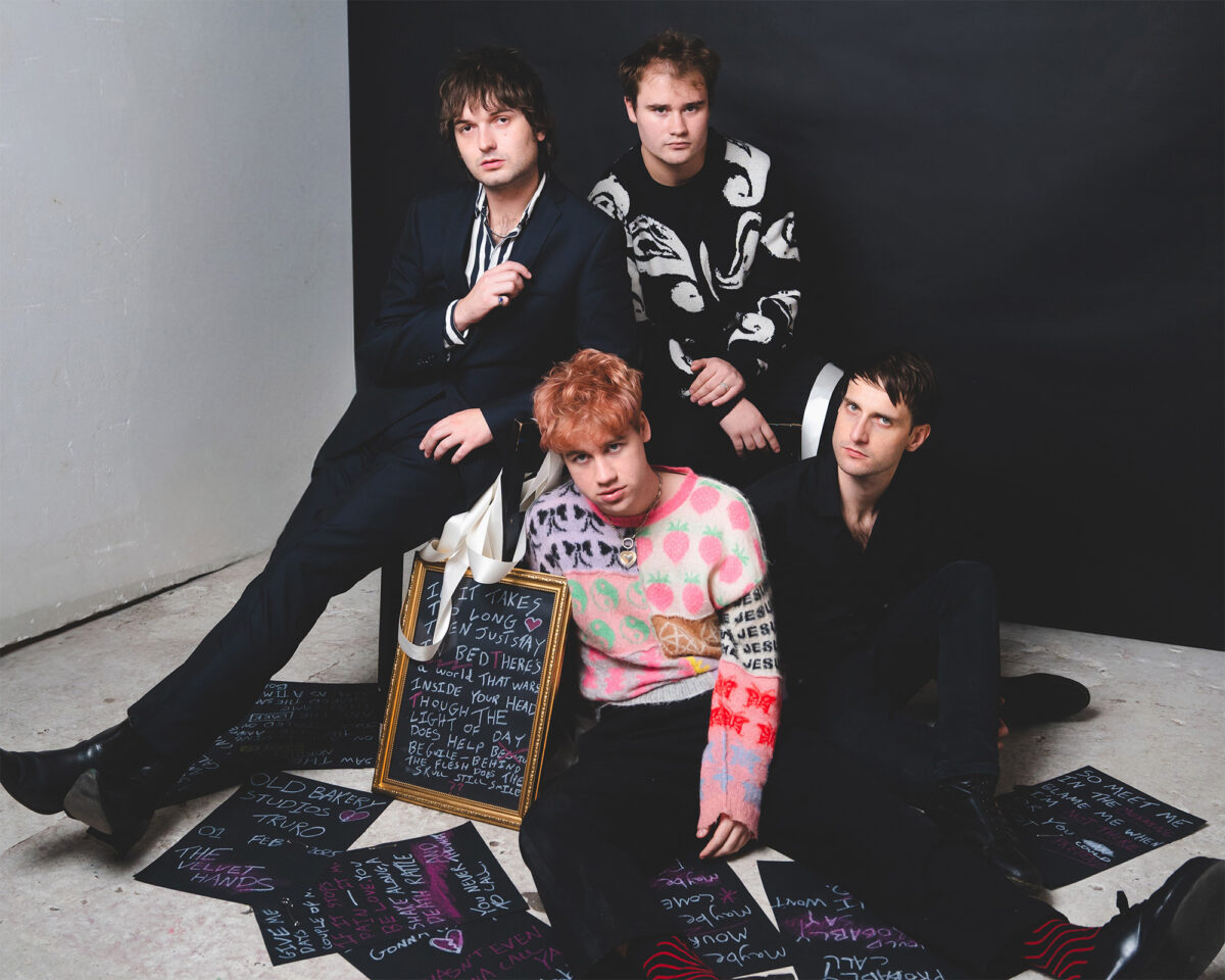 A professional studio portrait of a four-person band. They're positioned in a simple white and black corner set with a minimalist aesthetic.
