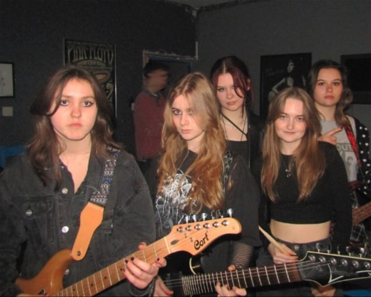 An all-female band or music group posing in what appears to be a rehearsal room or music venue. In the foreground are four young women, three of whom are holding guitars.