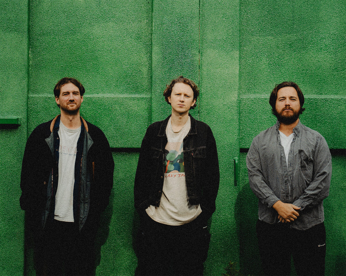 Three men standing in a row against a bright green textured wall or backdrop. They appear to be a music group posing for a promotional photograph.